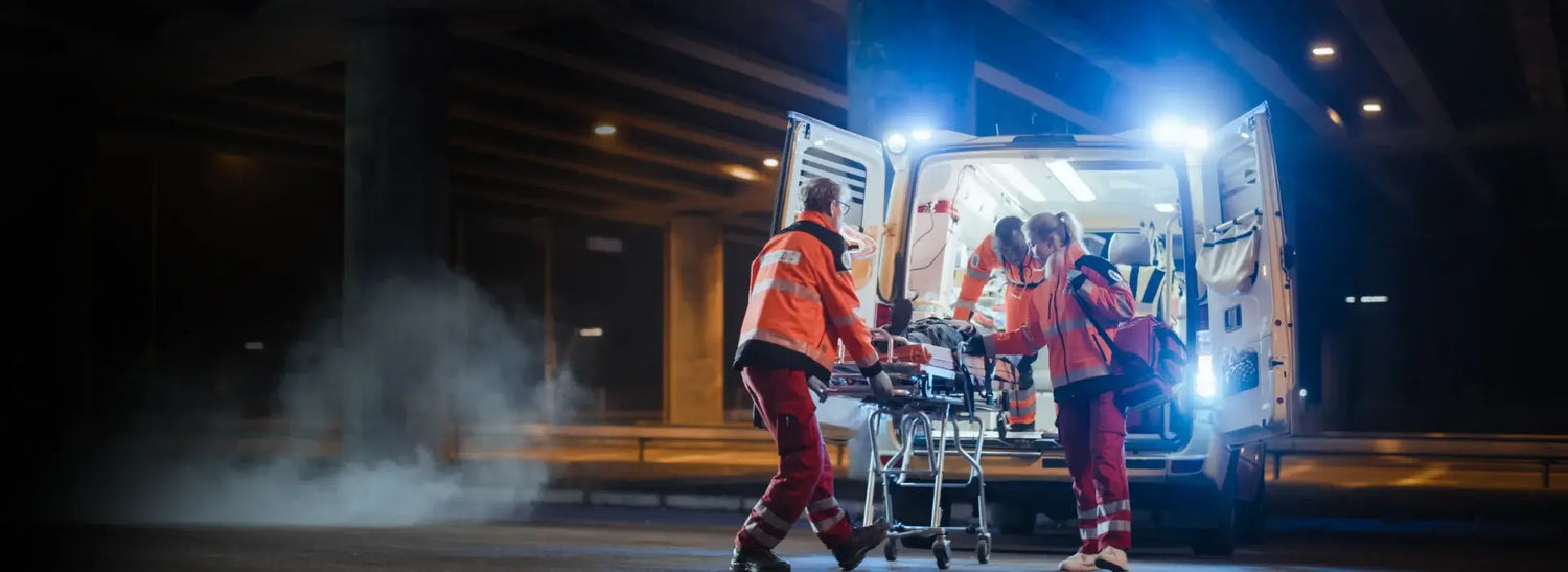 Two paramedics in orange uniforms loading a stretcher into an ambulance at night.